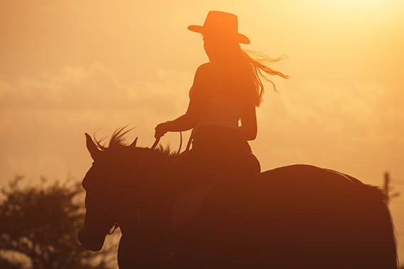 Sunset golden silhouette of young cowgirl in hat riding her horse